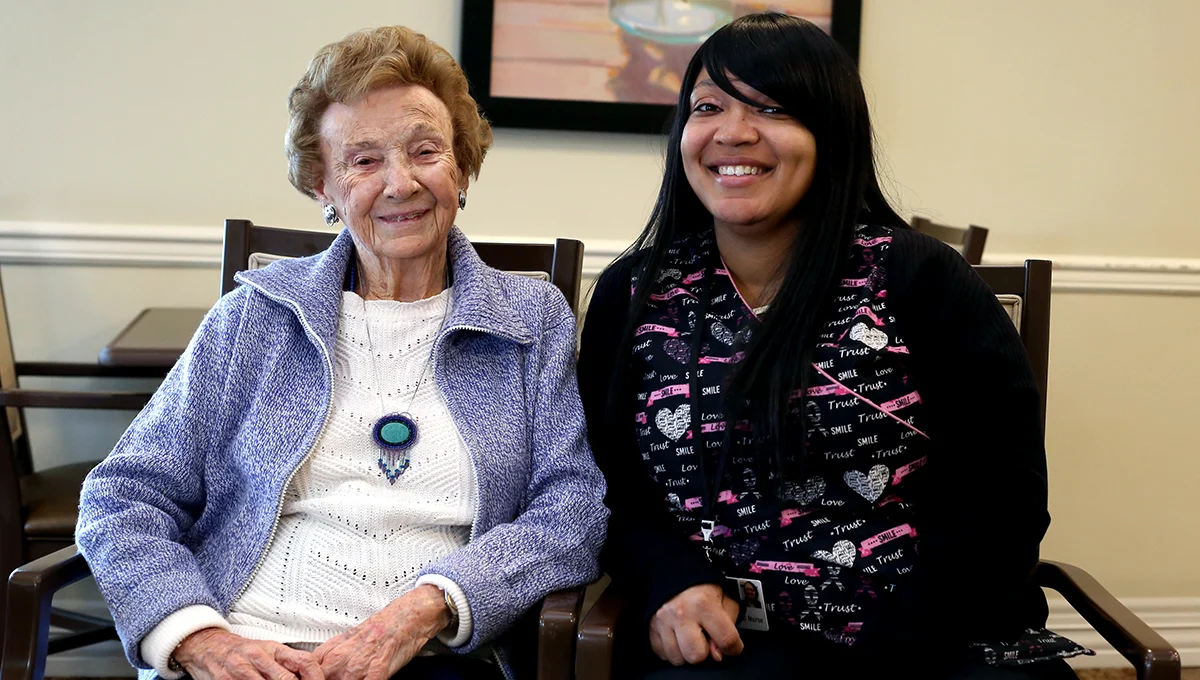 A smiling senior woman seated next to smiling nurse indoors.