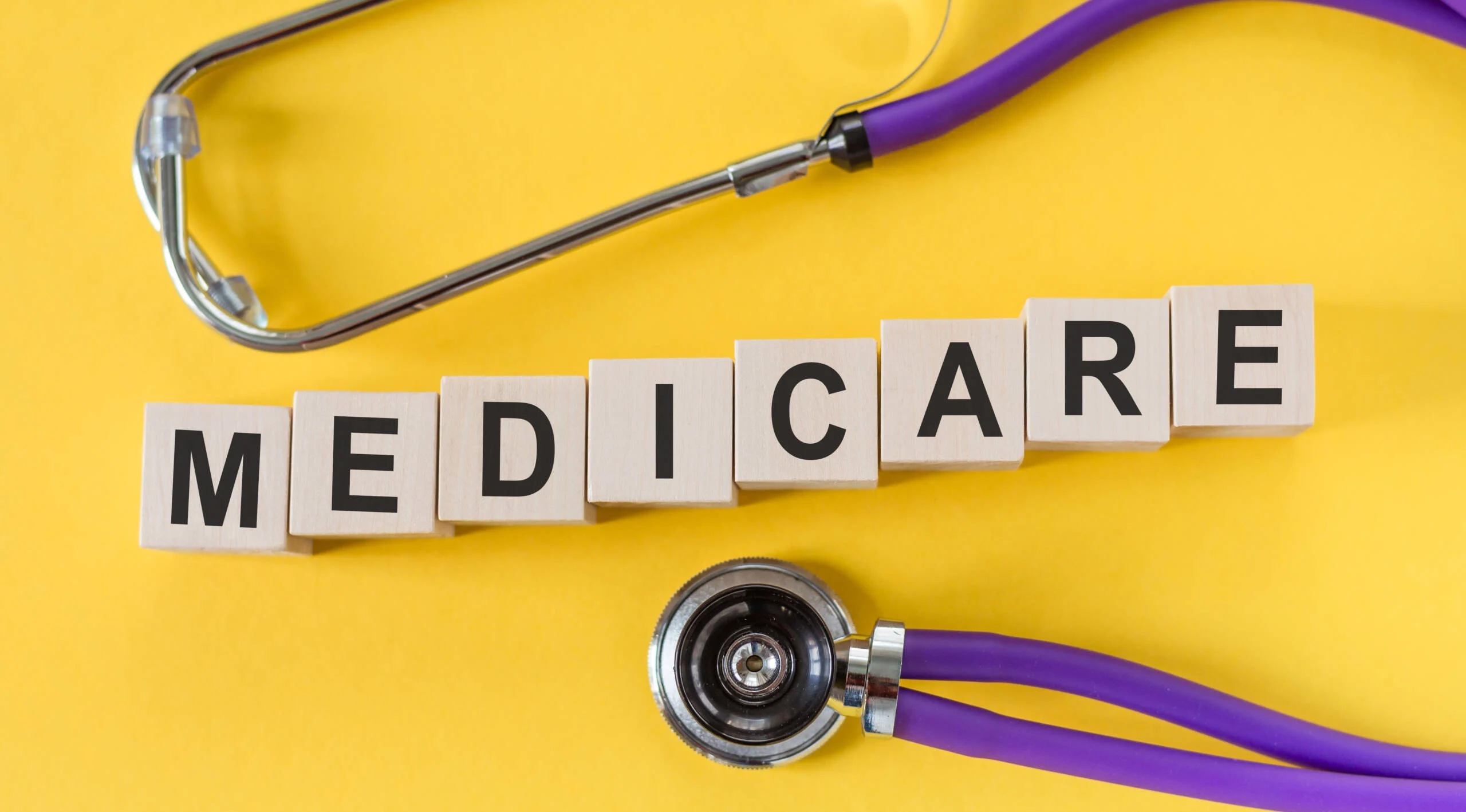 Medicare word written on cube shape wooden blocks on yellow table with stethoscope.