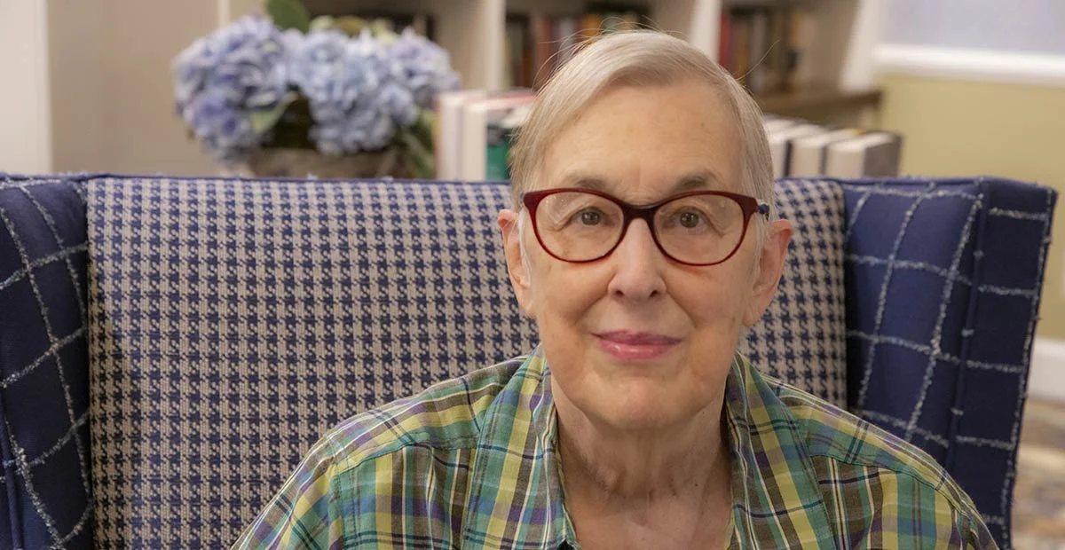 Smiling senior woman with glasses seated in a blue chair in a library.