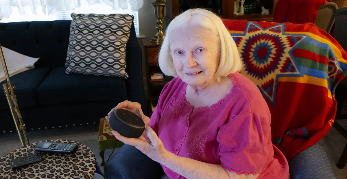 Smiling senior woman seated in her apartment holding out an Echo Dot.