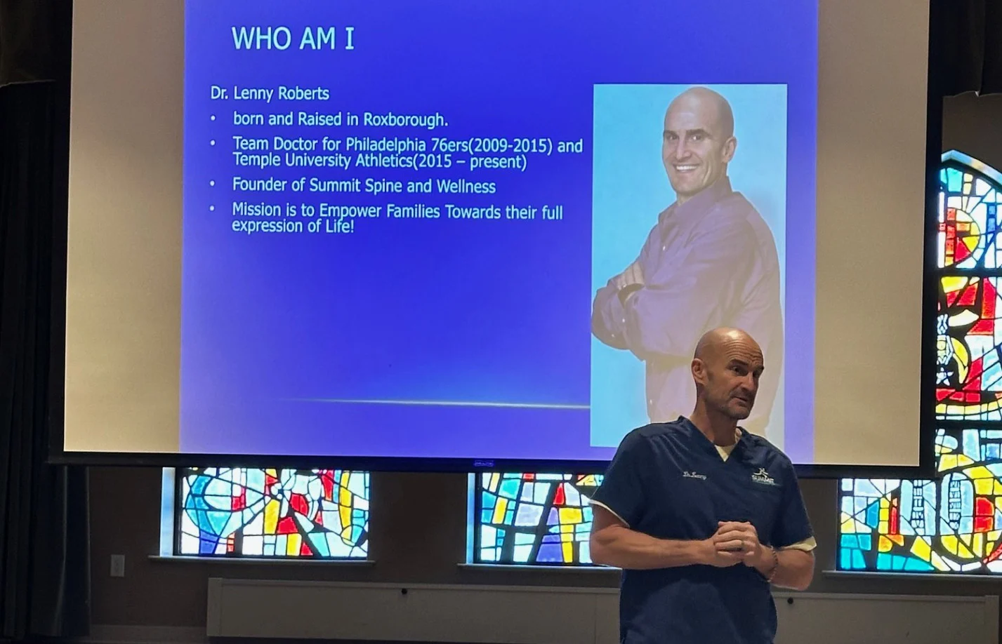 Man dressed in scrubs standing in front of slideshow presentation as he presents to a group.
