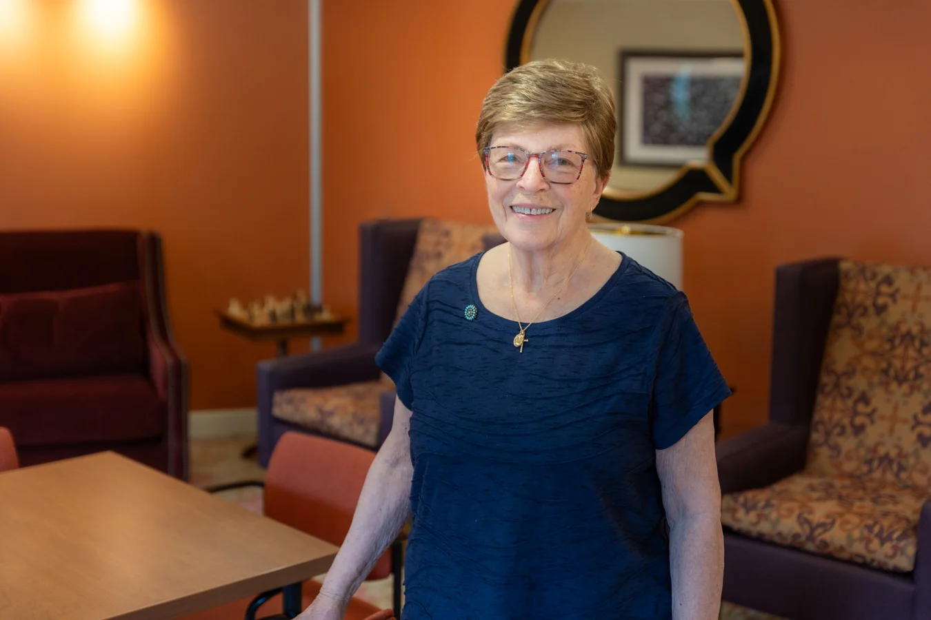 Smiling senior woman standing in lobby with chairs and a mirror behind her.