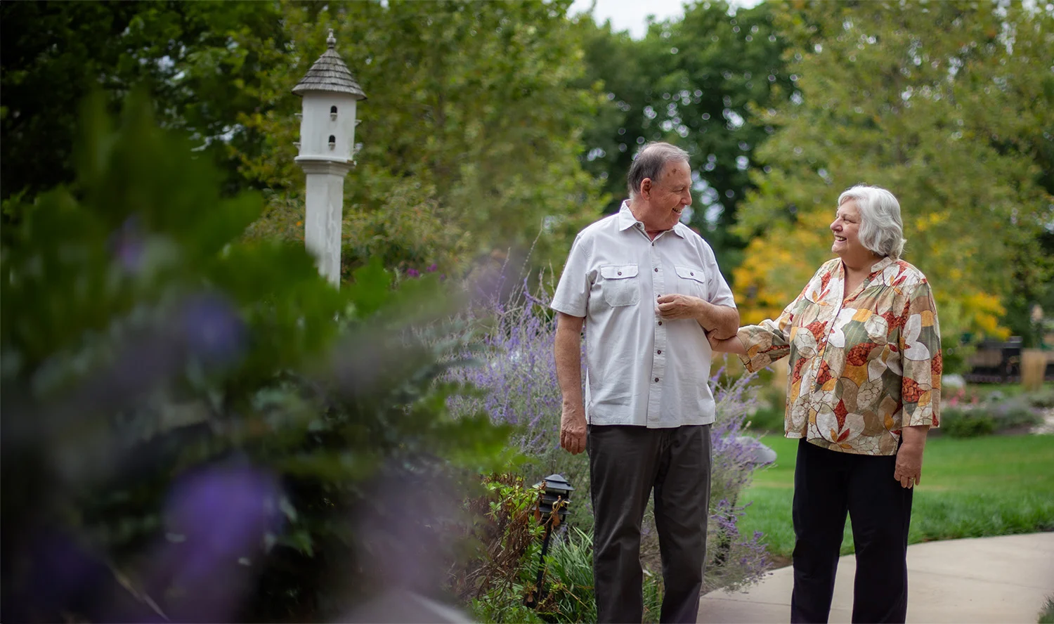 Smiling senior couple arm-in-arm walking on trail surrounded by flowers.