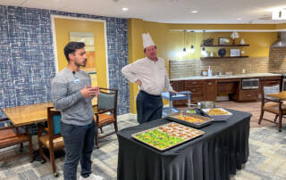 Dietician and chef standing in front of room with table set with healthy food samples