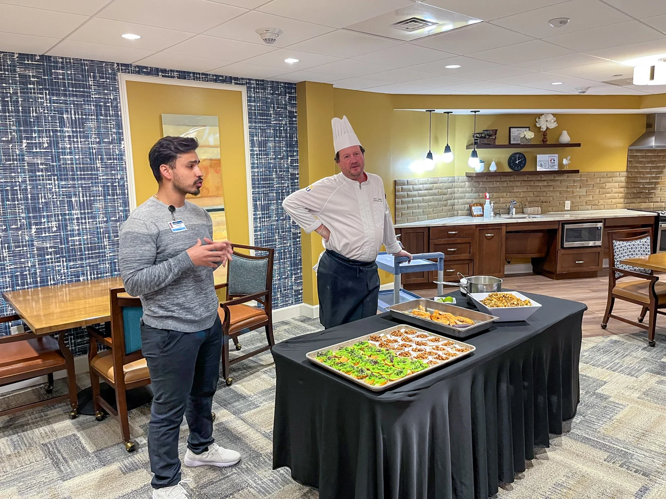 Dietician and chef standing in front of room with table set with healthy food samples.