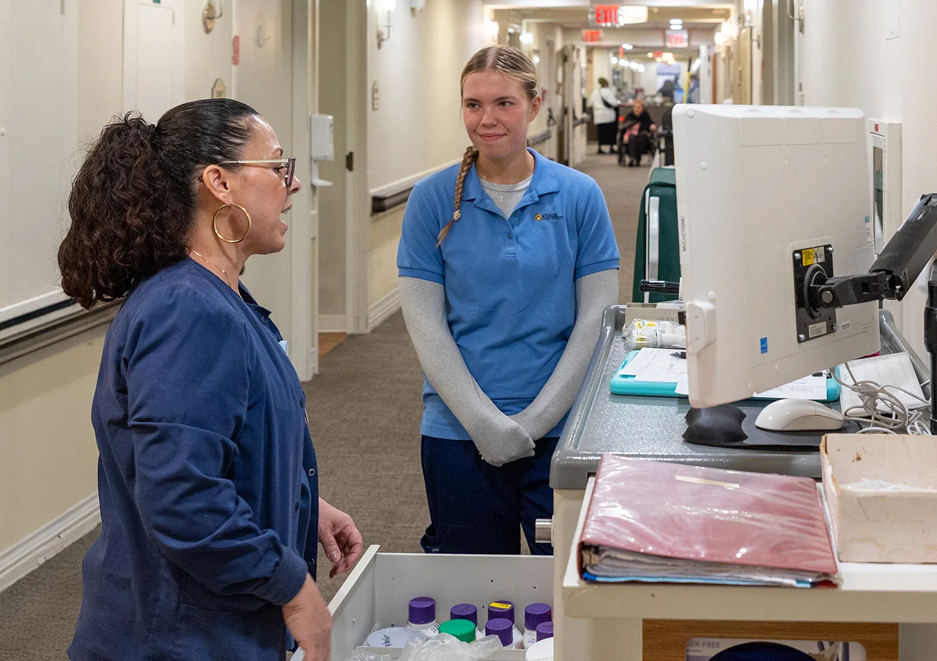 Nursing student and nurse speaking in hallway by a station with a computer.