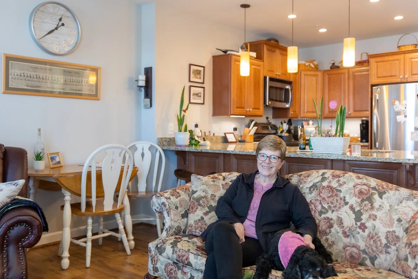 Smiling senior woman sitting with her black dog on floral couch in a well-lit apartment.