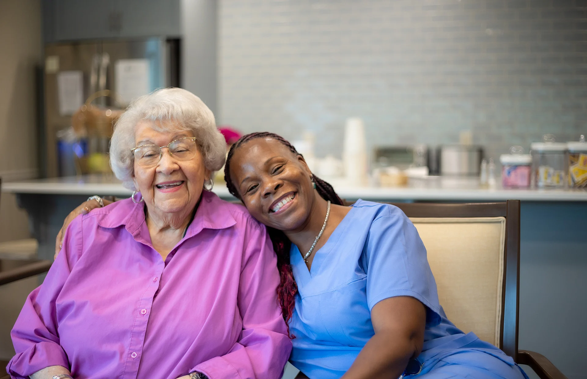 Smiling senior woman sitting with a smiling nurse, who has her arm around her.