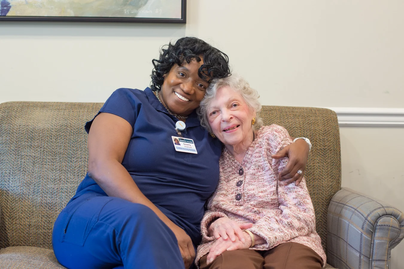 Senior woman and nursing assistant. A nurse hugging a smiling senior woman while the two are seated on a couch.