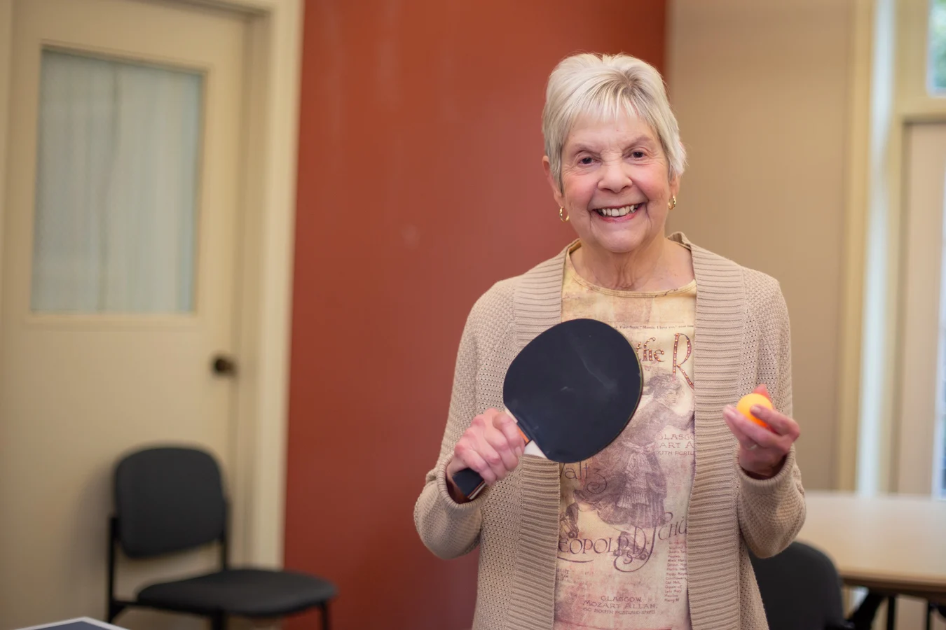 Smiling senior woman holding up a paddle and ping pong ball while standing indoors.
