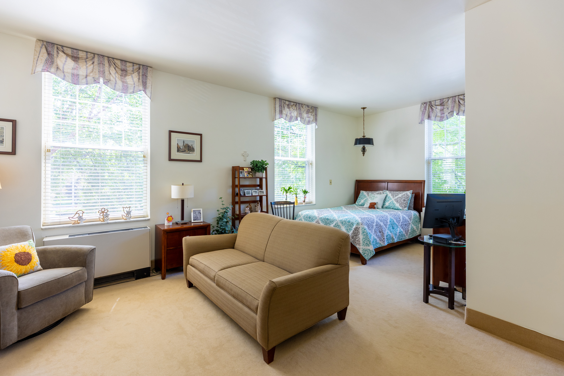 Wide shot of a well-decorated, brightly-lit room with a bed, dresser, bookshelf and couch in a personal care room.