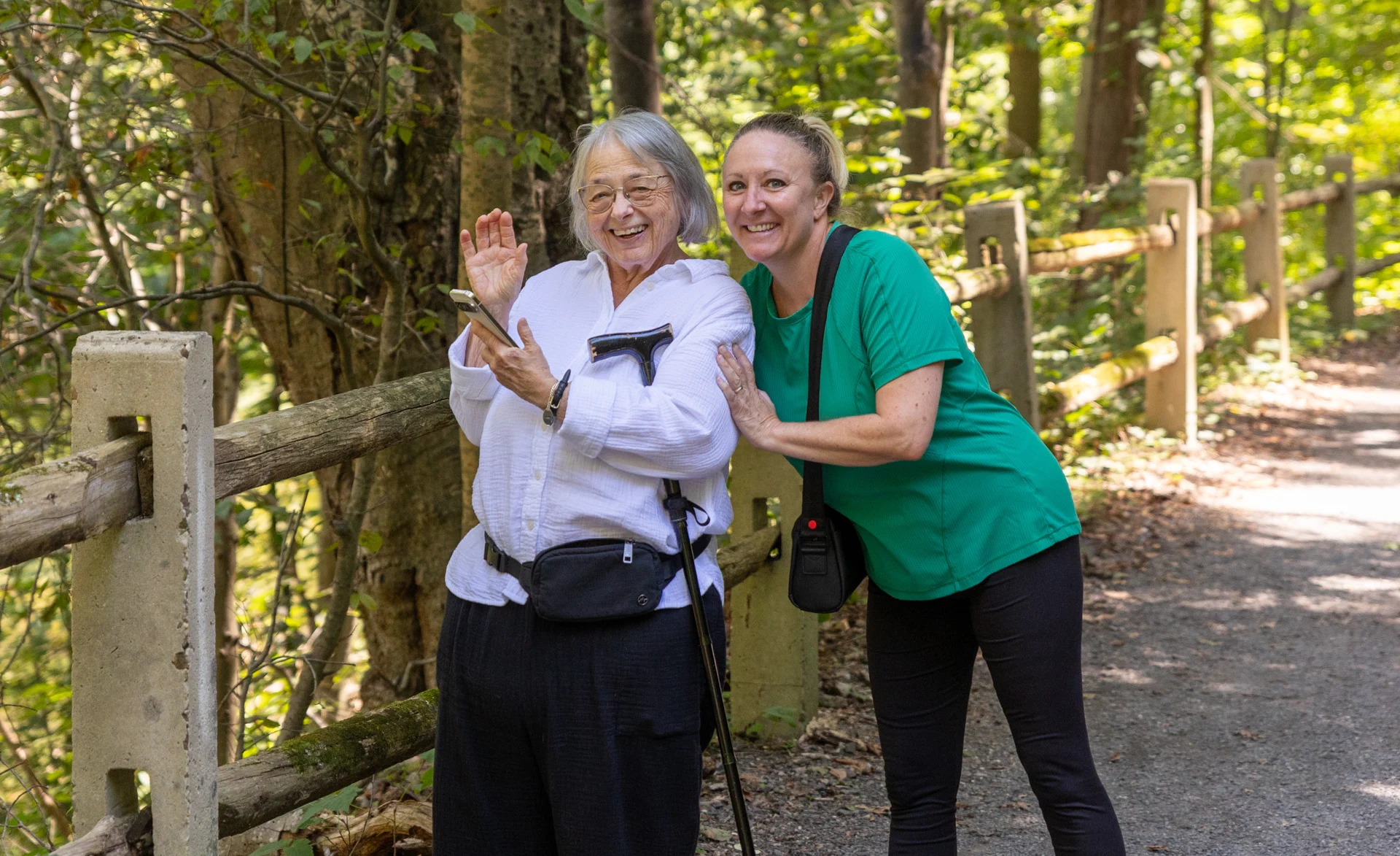 Senior woman waving while posed with staff member. Senior woman smiling and waving, standing next to a smiling staff member outdoors.